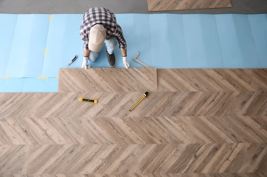 Ouvrier installant un parquet chevron en int&eacute;rieur, agenouill&eacute; sur sous-couche bleue, manipulant lames de parquet stratifi&eacute; ou contrecoll&eacute;