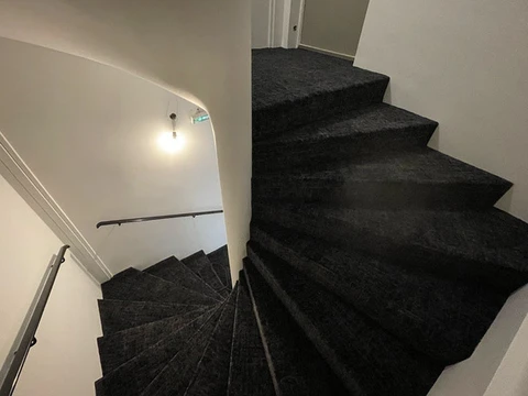Escalier intérieur en colimaçon avec marches revêtues de moquette noire, rampe métallique noire fixée au mur blanc